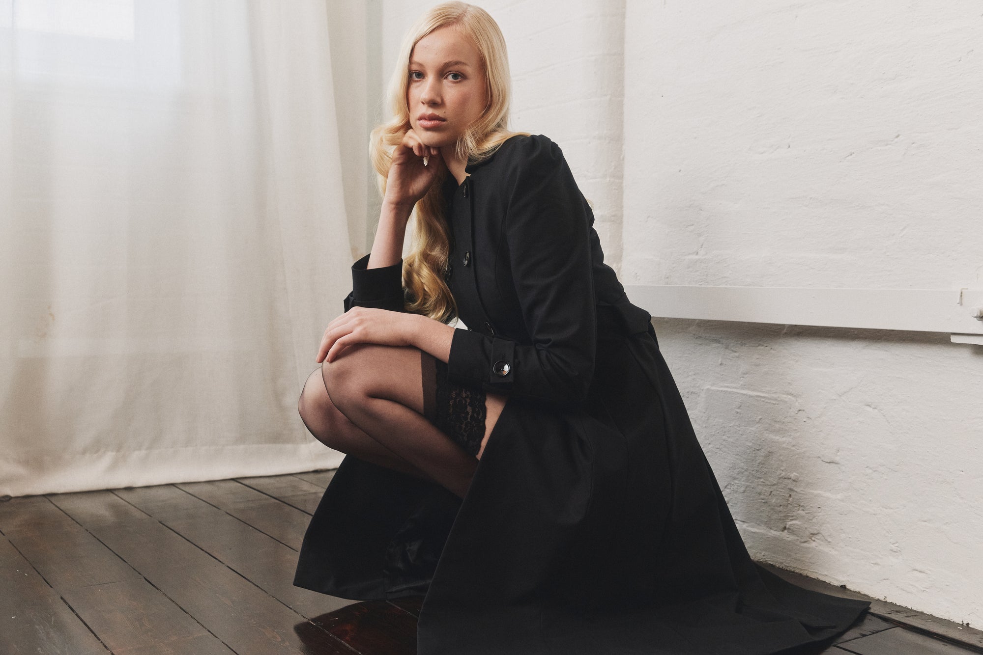 Woman in a black coat sitting on the floor against a white wall.