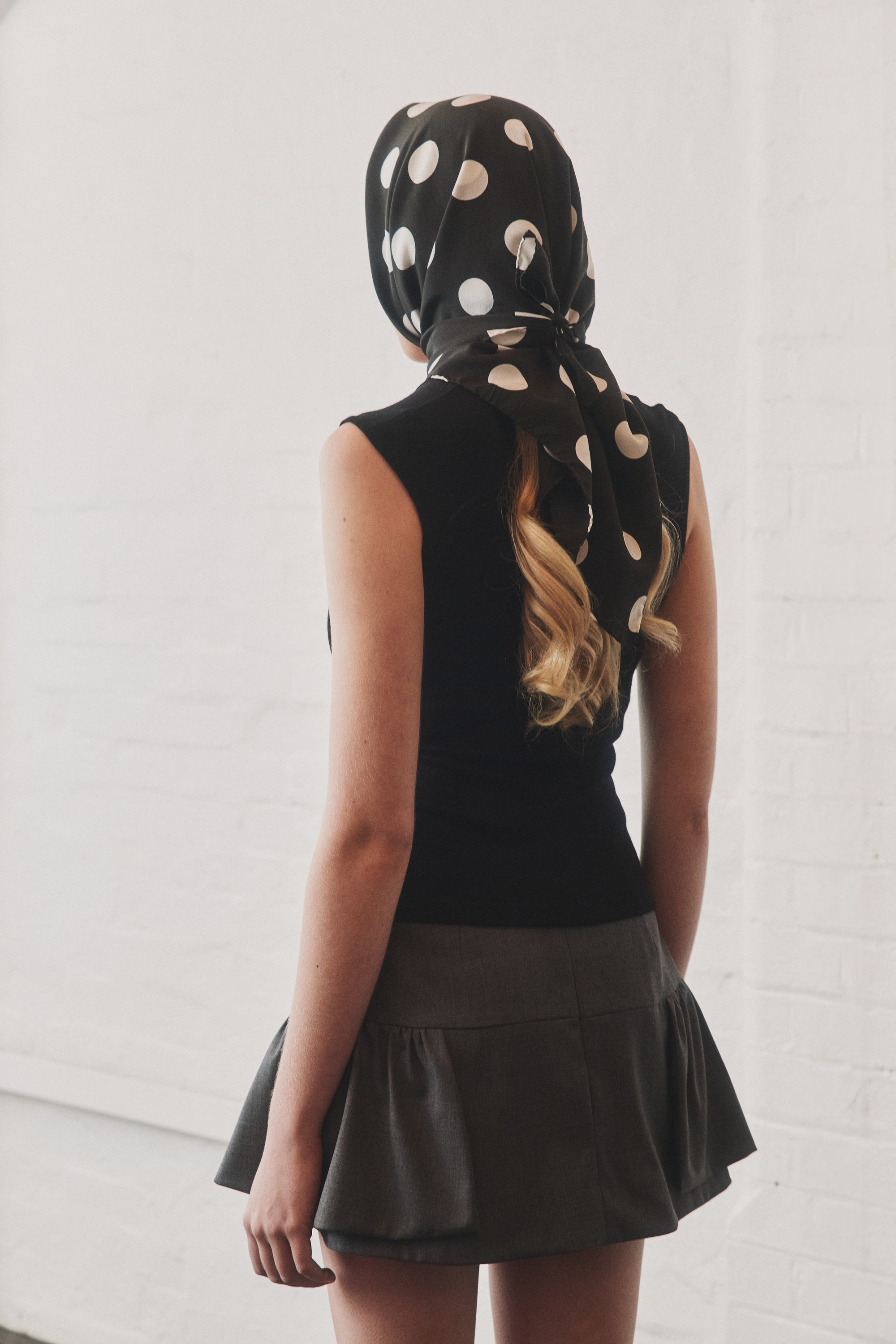 Woman wearing a black silk polka dot scarf by Australian label nessi tied over the head, photographed from the back against a white brick wall.