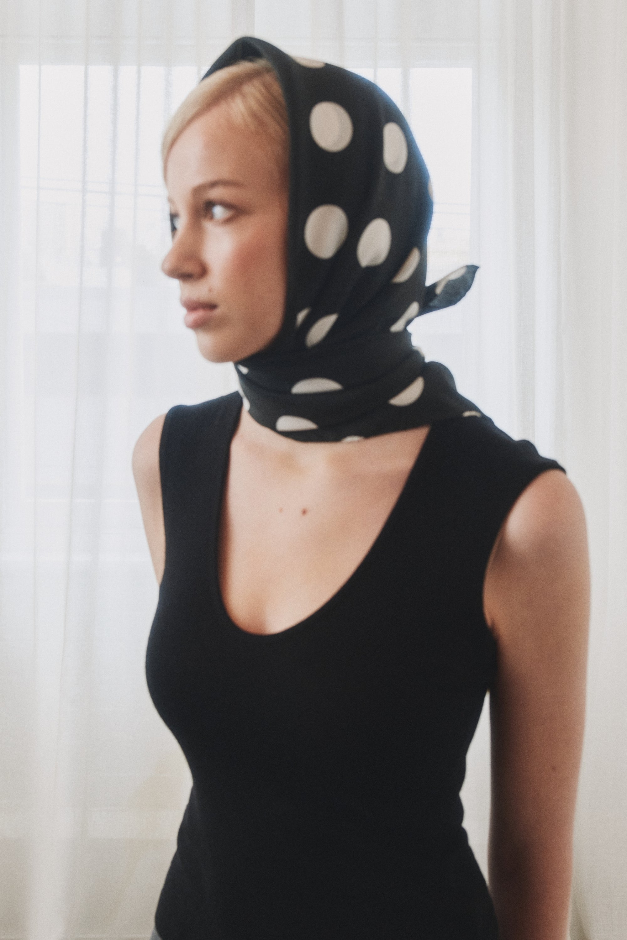 Woman wearing a black and white polka dot silk scarf by Australian label nessi, styled over the head against a soft white curtain backdrop.