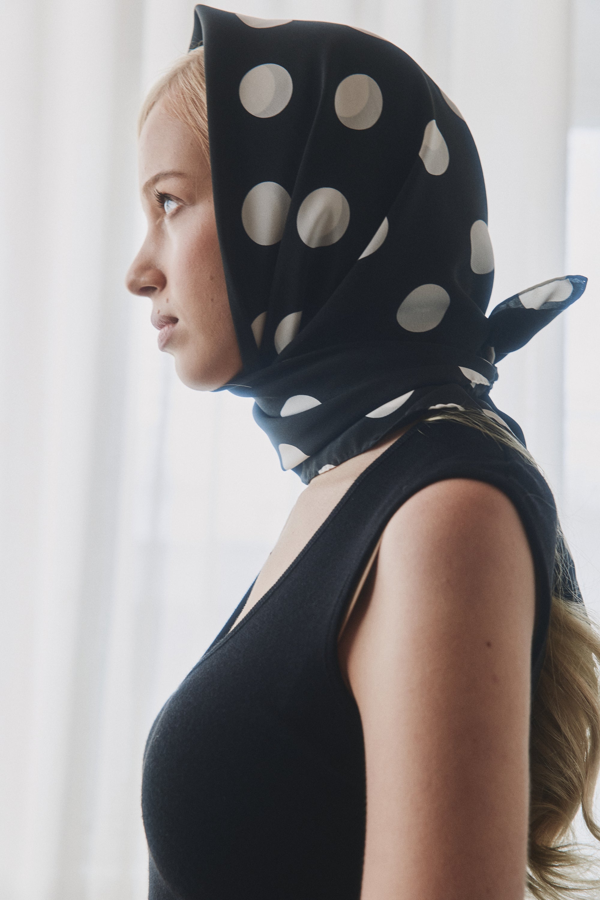 Woman wearing a black and white polka dot silk scarf by Australian label nessi, styled over the head against a soft white curtain backdrop.